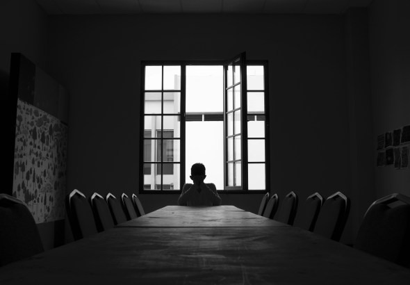 Silhouette of a person sitting alone at a long table in a dark room, with an open window behind them letting light in.