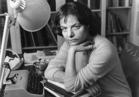 Woman at a desk with a vintage typewriter, resting her chin on her hands among books and papers in a cluttered study.