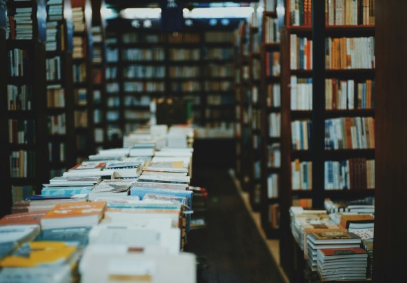 Bookstore aisle with a long table of stacked books in the foreground and tall shelves along the walls in the background, dimly lit.