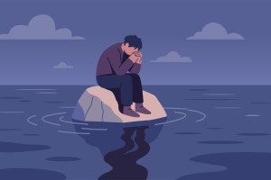 A person sits on a rock by the water, head in hands, appearing sad or distressed at dusk.