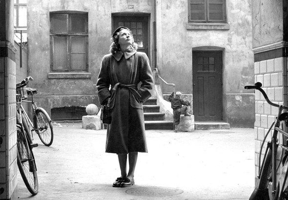 Woman in a belted coat stands in a narrow alley between old buildings, looking upward; bicycles lean nearby.