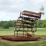 Rusty sculpture made from stacked chairs on a circular platform in a park, with a white abstract form on top.