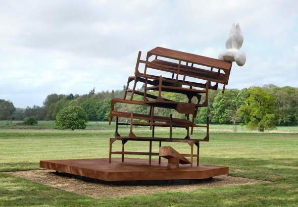 Rusty sculpture made from stacked chairs on a circular platform in a park, with a white abstract form on top.