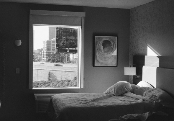 Dimly lit hotel bedroom with an unmade bed in the foreground and a window showing a city street outside.
